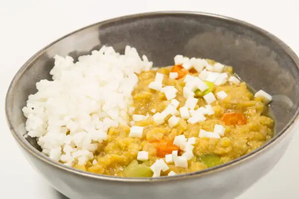 Un tazón de arroz blanco junto a un guiso cremoso de lentejas rojas con trozos de queso y verduras.