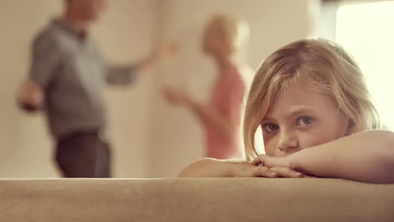 Niña asustada, escondida tras un sofá, mientras sus padres discuten delante de los hijos.